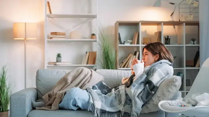 Woman sat up on a sofa with a blanket drinking a hot drink